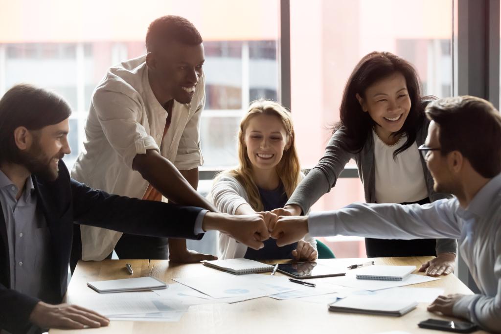 Happy,Diverse,Employees,Team,Joining,Fists,,Sitting,At,Table,In